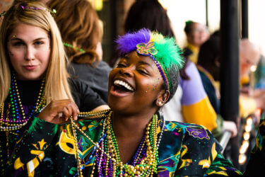 Woman wearing a feathered headband smiles and shows her gold, green and purple Mardi Gras beads.